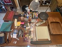 Full overview of the office supplies spread on wooden table showing all items in the lot including lamp, paper cutter, stapler, scissors, pens, glass apple, organizers, and more.