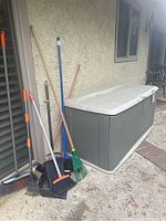 Photo showing large gray Rubbermaid outdoor storage box placed against a wall with various outdoor brooms and rakes leaning beside it.