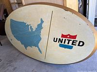 Full view of the large oval hand painted United Airlines wall sign against garage background. Shows the blue map of the US with route lines on the left, and the United Airlines logo on the right.