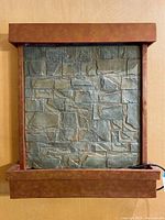 Front view of the wall hanging water fountain showing the carved stone brick pattern and rust-colored frame.