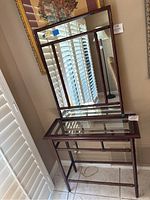 Cherry wood vanity console table with a glass top and lower wooden slat shelf, paired with a large mirror that has a carved bamboo-style wood frame. Both items are shown together near window blinds.