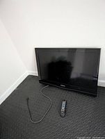 Front view of 32 inch Samsung TV resting on floor with power cord and remote control in front, showing some light scratches and no feet attached.