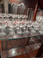 Set of 12 etched crystal champagne glasses displayed on glass shelf inside a wooden cabinet.