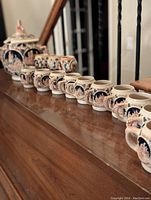 Full line-up of ten small mugs and the large cookie jar with lid, showing the detailed decorative images and shape, displayed on wood surface.