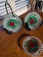 Two iron stands holding five and three glass coasters with scalloped edges and rose design, one coaster laid flat on wooden surface.