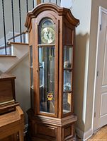Full view of the tall wooden grandfather clock with glass doors and side shelves displaying small sculptures.