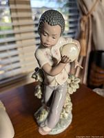 View of porcelain figurine facing front showing boy holding drum, textured curly hair, floral shirt, and pedestal with flowers