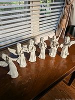 Set of 6 white porcelain angel figurines arranged on a wooden surface, showing the full choir/set looking towards the sunlight.