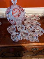 Punchbowl and 12 matching mugs arranged on wooden cabinet showing intricate red bird crystal design.
