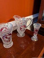 Wide view of three Nachtmann crystal items with red bird motif on wood surface featuring clear, deep red heart-shaped accent and detailed cut crystal patterns.