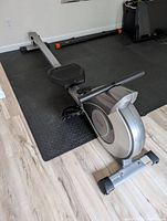 Full side view of the silver and black rowing machine placed on a black rubber gym mat on light wood flooring.