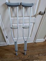 Pair of silver aluminum adult crutches with grey padded underarm supports and hand grips, standing against white door on wood floor.