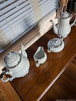 Coffee pot, teapot, creamer, and sugar bowl in white china with silver trim and black scroll accents on wood surface.