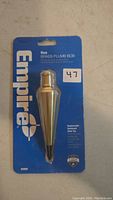 Photo of new Empire 8 oz brass plumb bob in original sealed blue packaging, showing front view with label and replaceable hardened steel tip visible.