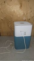 Front view of Blueair air purifier on concrete floor against wooden wall showing unit and power cord.