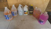 Seven cleaning product containers arranged on floor against wood backdrop. Products include assorted degreasers, disinfectant, mold control, siding and masonry cleaner, bug and sap cleaner. Colors vary from orange, white, dark yellow-black, red, to purple. Some containers partially used, some unopened.