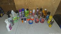 Overview of assorted aerosol sprays, cleaning fluids, and various tapes arranged on a table against a wooden wall.