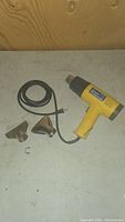 Photo showing yellow heat tool with attached power cord and three different metal nozzle attachments laid out on a flat surface with small retaining clip