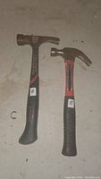 Photo showing two hammers on the floor, a larger Husky 20oz framing hammer and a smaller claw hammer with red and black handle.