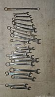 Top-down view of 20 combination wrenches arranged by size on a floor surface. Each wrench has an open-end and a box-end. Various metric sizes observable from small to large.