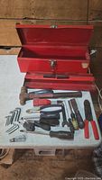 Open red metal toolbox with hand tools laid out in front on white surface, including hammer, screwdrivers, hex keys, wire cutters, utility knife and pliers.