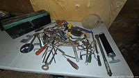 Wide view of a table filled with mixed hand tools, metal funnel, grease gun, and livestock feed bucket arrangement showing condition and number of tools.