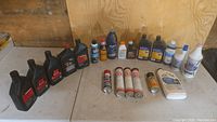 Lot of various automotive oils, lubricant bottles and spray cans arranged on the floor against plywood wall.