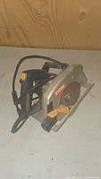 Side view of the Ryobi electric circular saw showing blade guard, handle, and power cord on a plain surface.