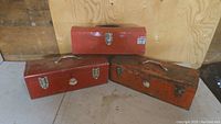 Photo showing three red metal tool boxes closed and arranged on floor against wood wall