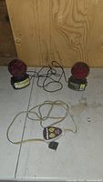 Photo of two red wireless towing lights with magnetic bases and attached wiring on white surface against plywood background.