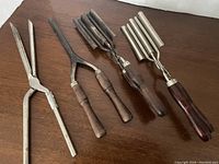 Five vintage hairstyling tools arranged on wooden surface showing different styles of tongs and barrel irons with wooden handles.