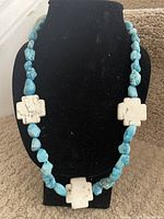 Front view of the 20 inch turquoise bead necklace on black display showing irregular turquoise stones and 3 white cross-shaped beads.