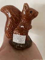 Front and side view of the ceramic squirrel decanter showing rich brown glaze and glossy finish. A handwritten label on the base describes the item.