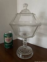 Front view of clear Early American Pressed Glass covered compote with lid, showing hexagonal shape and pedestal base next to soda can for scale.