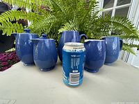 Front view of all five Moorcroft pottery pieces with a soda can for scale, showing two lidded pots and three open jugs in blue speckled glaze.