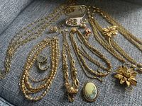 Overview of the assortment showing various gold-tone necklaces, earrings, brooches, a compact, and jewelry pieces on a gray fabric background