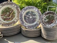 Stacks of dinner plates, salad/cake plates, and bowls with rural scenes and borders photographed outdoors with greenery background.