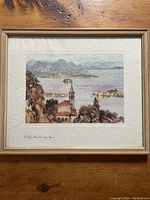Framed watercolour painting showing a lakeside scene with mountains and a church steeple in the foreground.