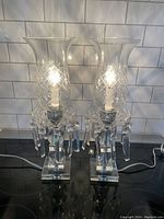 Pair of crystal table lamps lit, showing cut crystal globes with diamond pattern and rectangular prism accents on black counter with white tile background.