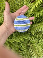 Photo showing an adult hand holding the bead-covered spherical ball against a green leafy background for size reference and craftsmanship detail.