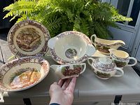 Photo showing full set of ceramic serving pieces with Royal Staffordshire England patterns laid out in natural light beside a fern plant.