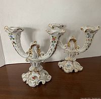 Pair of white porcelain candlesticks with blue, pink, green floral handpainted designs and gold accents on a wooden surface. Each has two arms and decorative bases.