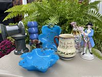 Composition of all lot items displayed on table with brick background and greenery, showing blue Moorcroft bowls, Motto Ware jug, Sitzendorf figurines, and antique turquoise glass piece