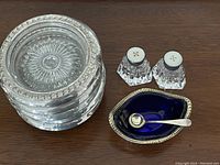 Overview of six sterling silver coasters stacked and pair of cut crystal salt and pepper shakers with white tops alongside a sterling silver salt cellar with cobalt blue liner and small sterling silver spoon.