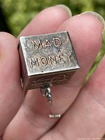Close-up photo of sterling silver cube pendant showing 'MAD MONEY' embossed on metal surface held between fingers.