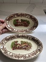 Top view of two oval serving plates showing rural scene and floral border
