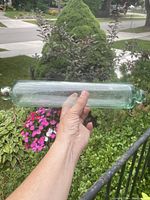 Full view of the hand blown glass rolling pin showing its length and clear light green tint, held horizontally outdoors.
