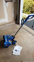 Full view of blue Earthwise electric snow thrower showing handle, wheels, and included user manual papers laid out on garage floor.