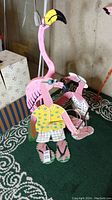 Three flamingo statues displayed on green patterned carpet: a tall wooden flamingo in pink with a yellow and black beak, and two smaller painted metal flamingos designed to hold plants, one dressed in a yellow shirt and shorts, the other with white spots.
