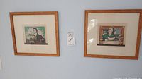 Photo of two framed artworks hung on a light blue wall, showing folk art of a farmer and a housewife.